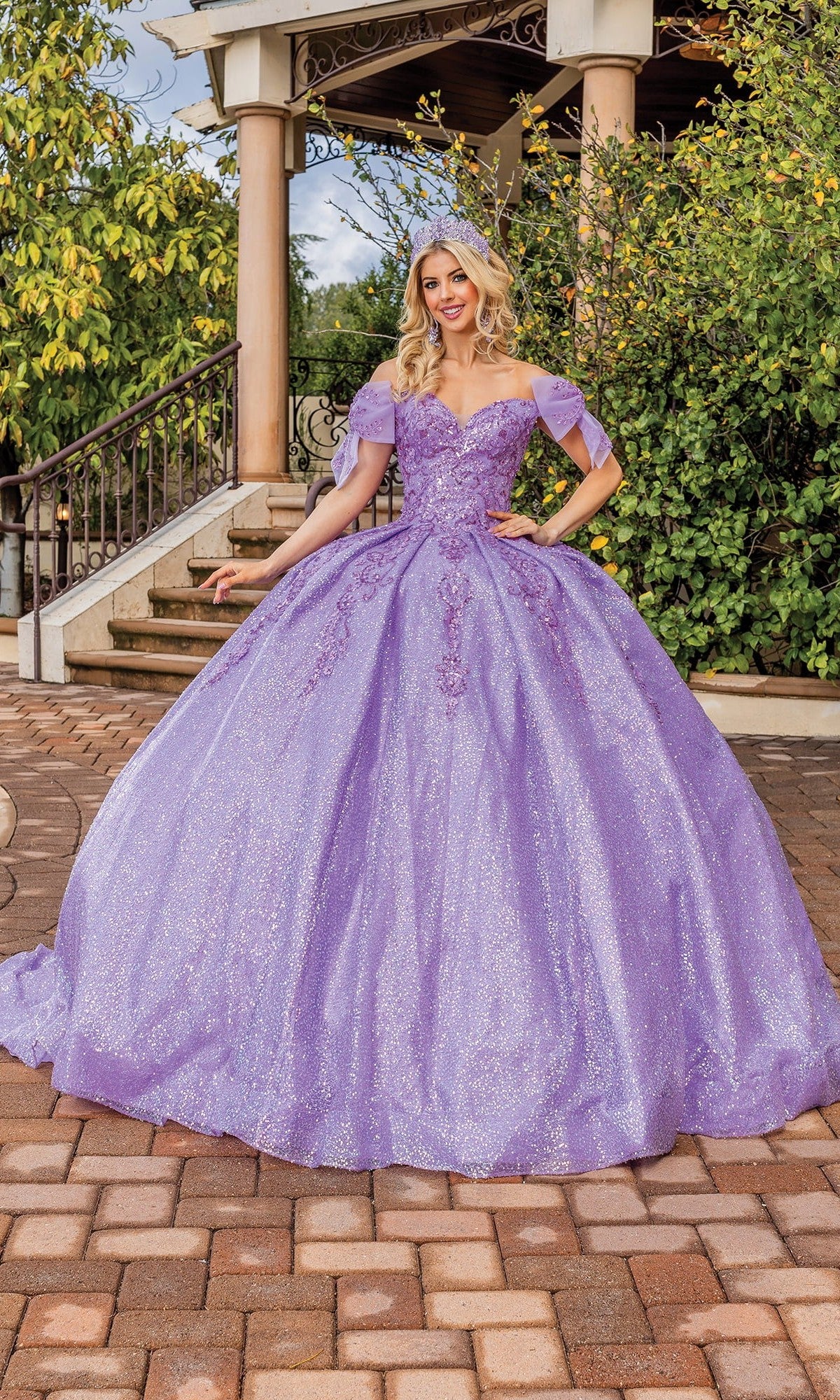 A woman stands outside posing gracefully in front of a gazebo and lush greenery, wearing the Quinceanera Dress 1804 By Dancing Queen—a sparkling lilac off-the-shoulder gown featuring floral appliqués and a sweetheart neckline.